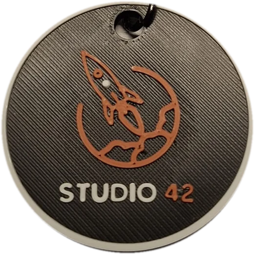 Llavero Studio 42 impreso en 3D: disco gris grafito con borde blanco, cohete y planeta en relieve cobre y chip NFC